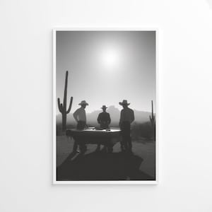 Op de afbeelding: Zwart-witfoto van drie figuren met cowboyhoeden rond een pooltafel in een woestijnlandschap. De zon schijnt fel, met cactussen op de achtergrond.