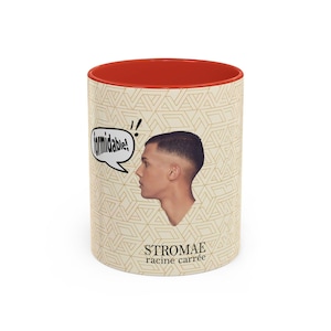 Może przedstawiać: Biały ceramiczny kubek z czerwonym wnętrzem. Kubek przedstawia profil mężczyzny, dymek z napisem "formidable!" oraz tekst "Stromae racine carrée" w beżowy geometryczny wzór.