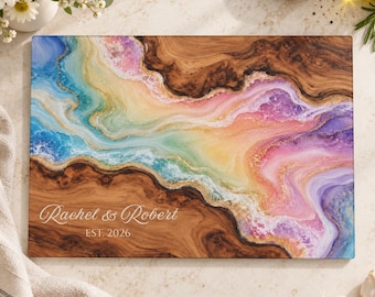 Peut inclure: Une planche à découper rectangulaire en bois avec une incrustation de résine tourbillonnante aux tons bleus, roses et dorés. Le grain du bois est visible. Le texte Rachel & Robert et EST. 2026 est gravé en blanc dans le coin inférieur gauche.
