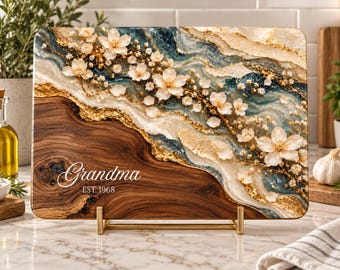 Peut inclure: Une planche à découper rectangulaire en bois avec un grain sombre d'un côté et un motif en résine bleu, blanc et doré de l'autre, ornée de fleurs blanches et du texte Grandma EST. 1968.