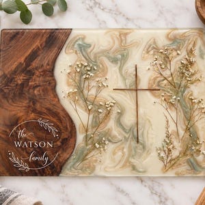 May include: A rectangular serving board featuring a dark wood section on the left and a cream, tan, and sage green marbled resin section on the right. The wood displays the white text "the WATSON family" inside a leaf wreath. Dried flowers and a thin metal cross are embedded in the resin.