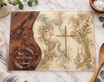 Peut inclure: Une planche de service rectangulaire avec une section en bois foncé à gauche et une section en résine marbrée crème, beige et vert sauge à droite. Le bois porte le texte blanc "the WATSON family" dans une couronne de feuilles. Des fleurs séchées et une fine croix en métal sont incrustées dans la résine.
