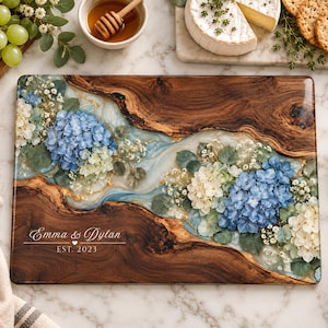 May include: A rectangular wooden serving board featuring a central river of clear resin embedded with blue and white hydrangea flowers and gold leaf accents. The board is engraved with the text Emma & Dylan EST. 2023 and has a natural wood grain.