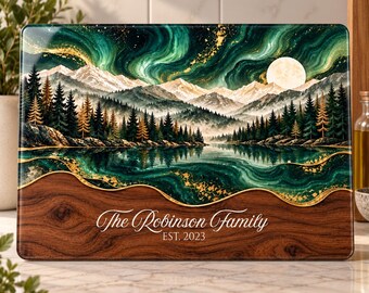 Peut inclure: Une planche à découper rectangulaire illustrant un paysage de montagnes enneigées, une forêt et une pleine lune sous un ciel d'aurore boréale verte. La partie inférieure présente un motif de grain de bois avec le texte The Robinson Family EST. 2023.