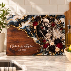 May include: A rectangular wooden cutting board featuring a dark blue and gold marble-effect resin inlay. The design includes a skeleton couple in wedding attire surrounded by red and white flowers, with the text Sarah & Dylan EST. 2024.