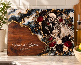 Peut inclure: Une planche à découper rectangulaire en bois avec une incrustation de résine effet marbre bleu foncé et doré. Le motif représente un couple de squelettes en tenue de mariage entouré de fleurs rouges et blanches, avec le texte Sarah & Dylan EST. 2024.