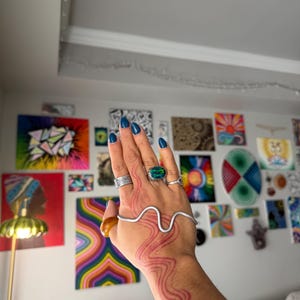 May include: A hand adorned with rings, a silver bracelet, and a red wavy design on the skin. The nails are painted a dark blue. The background features colorful abstract paintings.
