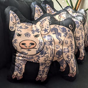 May include: Pig-shaped decorative pillows with a tattoo design. The pillows feature a light tan pig with a pink snout and black tattoos. The tattoos include stars, hearts, and the words "AMY LO INK". The pillows have black backs.