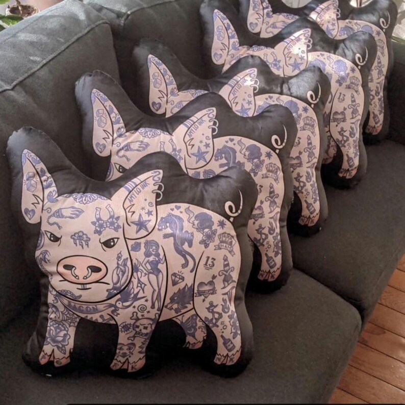 May include: A row of pig-shaped throw pillows with black bodies and light pink faces. The pigs are covered in blue tattoo-style designs. The pillows are arranged on a dark gray couch.
