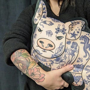 May include: A plush pig-shaped pillow with blue tattoo-style illustrations. The pillow features a pig face with a pink snout and ears. The tattoos include a banner that says "BACON" and the text "AMY LO INK".