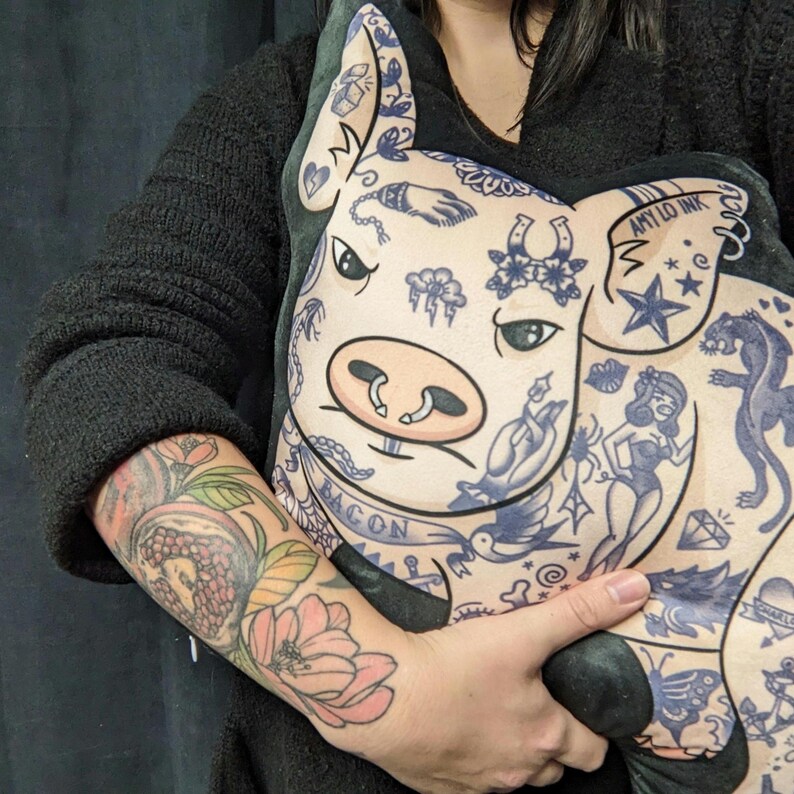 May include: A plush pig-shaped pillow with blue tattoo-style illustrations. The pillow features a pig face with a pink snout and ears. The tattoos include a banner that says "BACON" and the text "AMY LO INK".