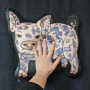 May include: A plush pig-shaped pillow with a light pink face and body, featuring blue tattoo-style designs. The pillow has a black border and includes the text "BACON" and "AMY LO INK".