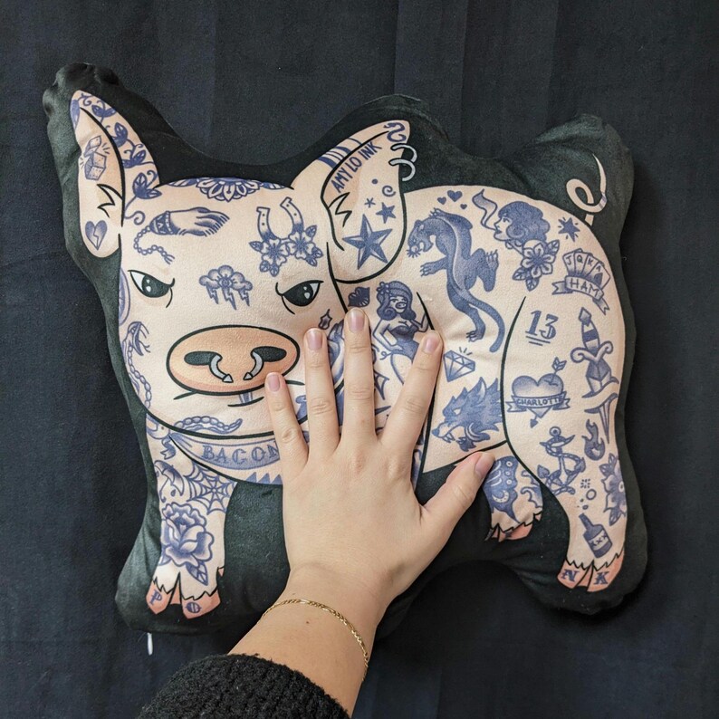 May include: A plush pig-shaped pillow with a light pink face and body, featuring blue tattoo-style designs. The pillow has a black border and includes the text "BACON" and "AMY LO INK".