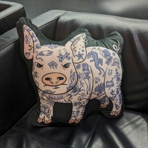 May include: A pig-shaped decorative pillow with a black border and a light pink face. The pig is covered in blue tattoos, including a horseshoe, a panther, and the word "BACON". The pillow is on a black leather couch.
