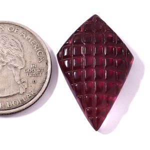 Könnte beinhalten: Ein facettierter, tiefroter Edelstein in Diamantenform. Der Edelstein hat ein gitterartiges Muster und liegt neben einer Münze zum Größenvergleich. Der Hintergrund ist weiß.