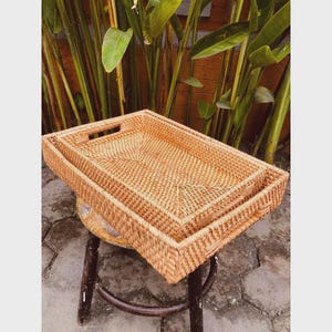 May include: Two rectangular woven rattan trays, one nested inside the other, sit on a small, round wooden stool. The trays are a light brown colour and have a textured surface. The background features green plants.