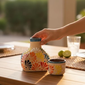 May include: A beige ceramic vase and small cup set with colorful floral designs in orange, yellow, blue, and white. The vase has a blue rim. The set is on a wooden table.