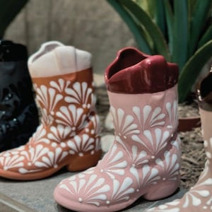 May include: A collection of ceramic cowboy boot planters in various colors, including black, pink, and brown. Each boot is adorned with a white floral pattern. The boots are displayed on a tiled surface with green plants in the background.