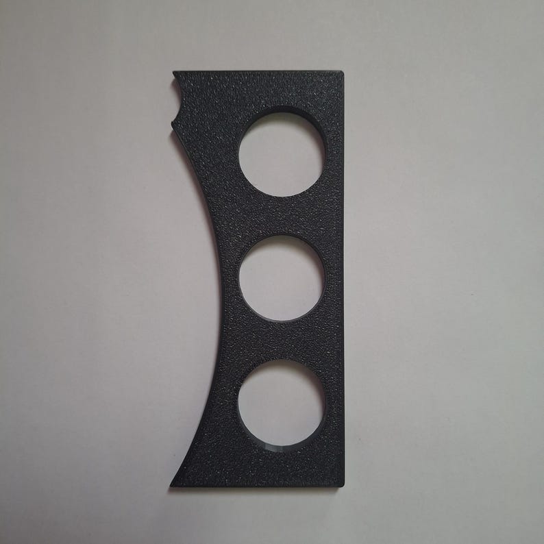 May include: A dark grey, textured, rectangular component featuring three circular cutouts arranged vertically in the centre. The left side of the object has a concave, curved edge, while the right side remains straight and flat.