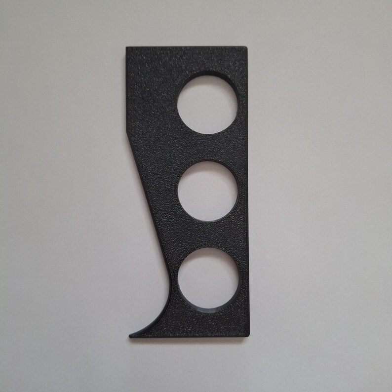 May include: A black, textured, flat, rectangular object with a tapered side and three circular cutouts arranged vertically. The item features a straight edge on one side and a curved, angled edge on the opposite side, creating a distinct geometric shape.