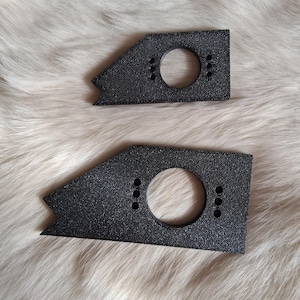 May include: Two black, textured metal brackets. Each bracket has a circular hole and three smaller holes. The brackets have a triangular shape with a notched end. The brackets are displayed on a fluffy, white surface.