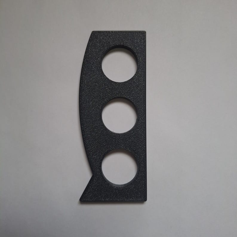 May include: A dark grey, textured, flat, rectangular object with a straight right edge and a curved left edge. It features three evenly spaced circular cutouts arranged vertically along the centre, with a small triangular protrusion at the bottom left.
