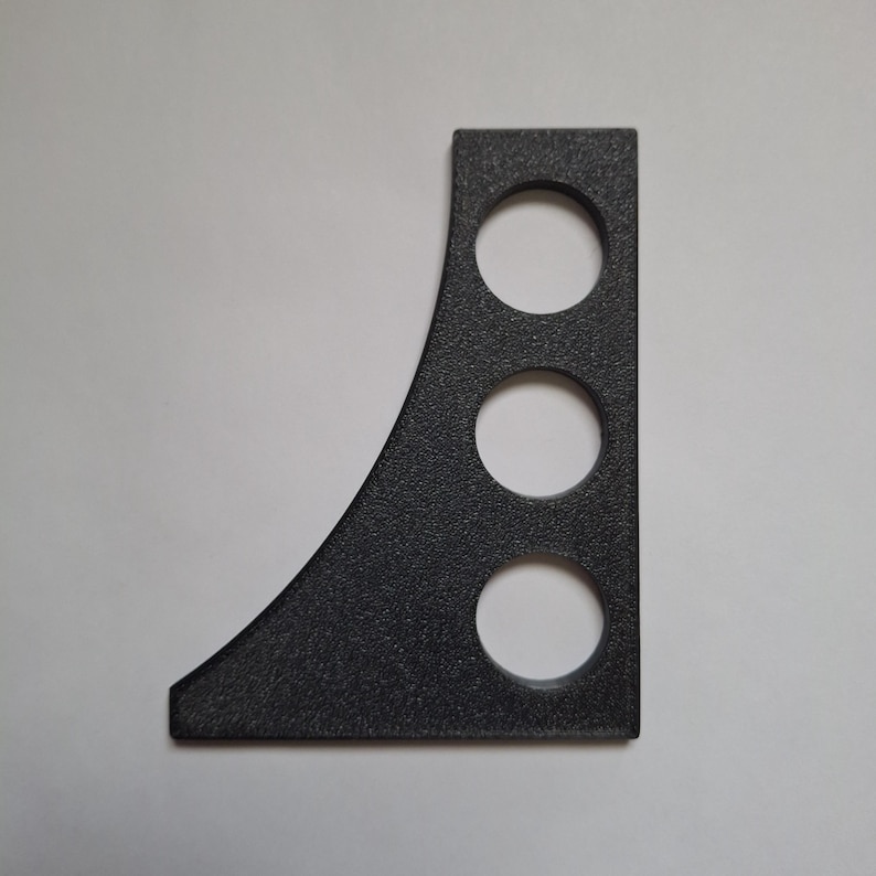 Op de afbeelding: Een platte, zwarte, getextureerde beugel met een rechte verticale rand en een gebogen zijrand. Het object is voorzien van drie cirkelvormige uitsparingen die in een verticale lijn zijn gerangschikt, wat een structureel onderdeel vormt.