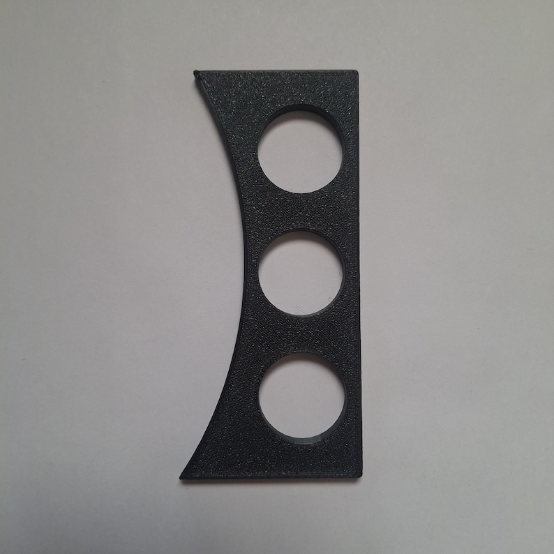 Puede incluir: Un componente rectangular de color gris oscuro con textura, que presenta un borde izquierdo curvo y tres recortes circulares dispuestos verticalmente en el centro. El objeto tiene un acabado mate y una textura superficial uniforme y granulada.