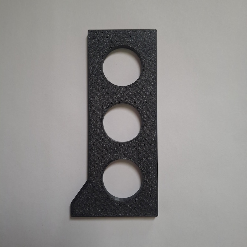 Puede incluir: Un objeto rectangular de color gris oscuro con una superficie texturizada que presenta tres recortes circulares dispuestos verticalmente. La esquina inferior izquierda del objeto est&aacute; inclinada, creando una forma geom&eacute;trica con una base plana.