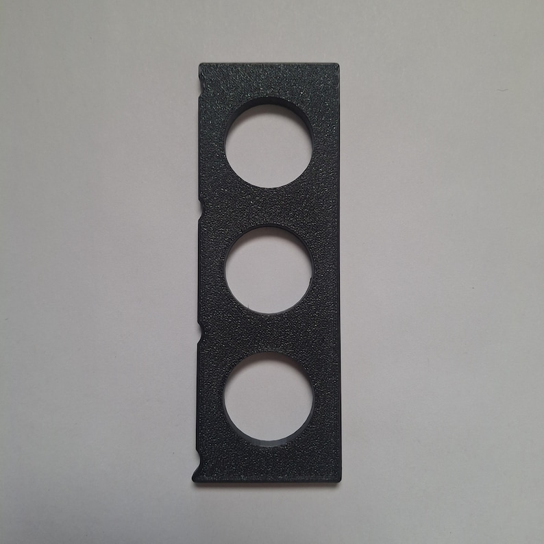 Pu&ograve; includere: Un oggetto rettangolare nero con superficie testurizzata, caratterizzato da tre fori circolari equidistanti disposti verticalmente. Il bordo sinistro dell'oggetto presenta tre piccole tacche semicircolari allineate alle aperture.