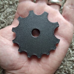Könnte beinhalten: Ein schwarzes, kreisförmiges, zahnradartiges Bauteil mit zehn ausgeprägten Zähnen am Außenrand und einem einzelnen, mittigen kreisförmigen Loch. Die Oberfläche des Objekts weist ein feines, lineares Texturmuster auf, das für 3D-Drucke typisch ist.
