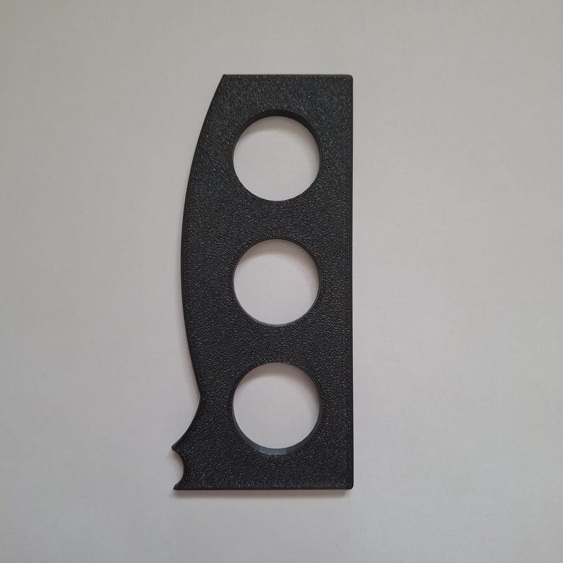 May include: A black, textured, rectangular tool featuring three circular cutouts arranged vertically in the center. The left side has a curved edge, while the bottom left corner includes a small, distinct semi-circular notch cut into the material.