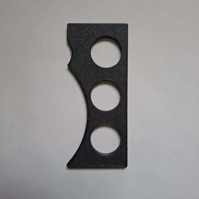 May include: A black, rectangular, textured tool featuring three circular cutouts arranged vertically along the centre and a curved, scalloped edge on the left side. The object has a flat, solid right edge and a matte, granular surface finish.