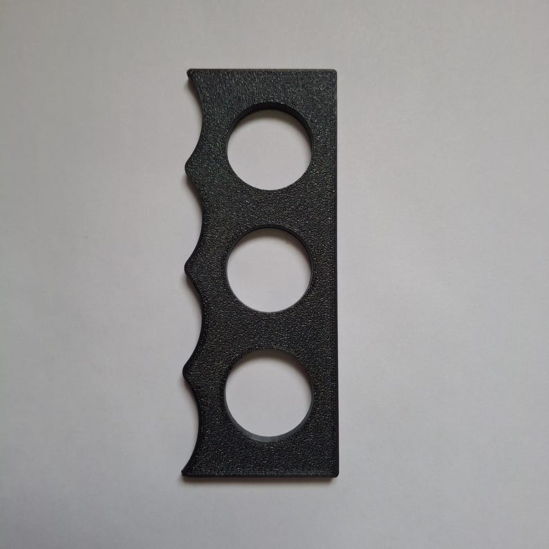 Puede incluir: Un objeto rectangular negro con una superficie texturizada que presenta tres recortes circulares dispuestos verticalmente en l&iacute;nea. Un lado del objeto tiene un borde festoneado con tres curvas distintas, mientras que el lado opuesto es recto y plano.