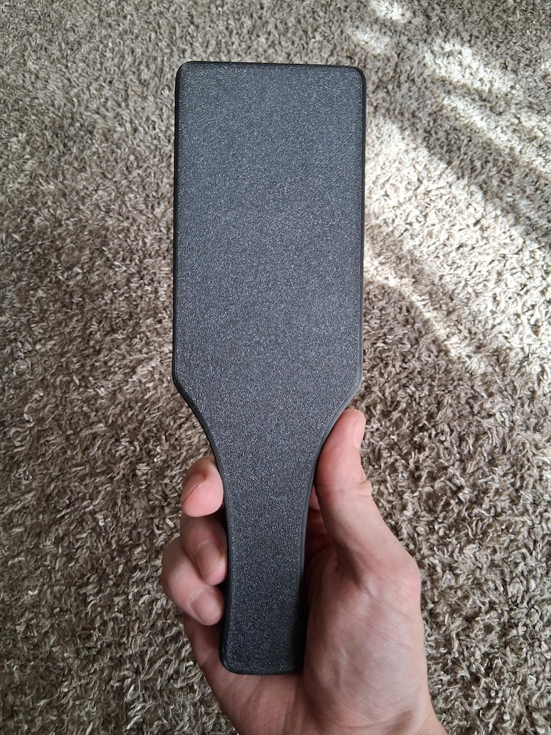 May include: A black paddle with a textured surface. The paddle has a rectangular head and a handle. The object is held in a hand against a neutral background.