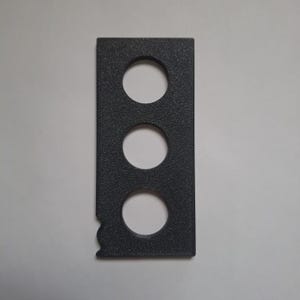 May include: A rectangular dark gray object with a textured surface featuring three evenly spaced circular cutouts arranged vertically in the center and a small, irregular notched indentation on the lower left side edge of the frame.