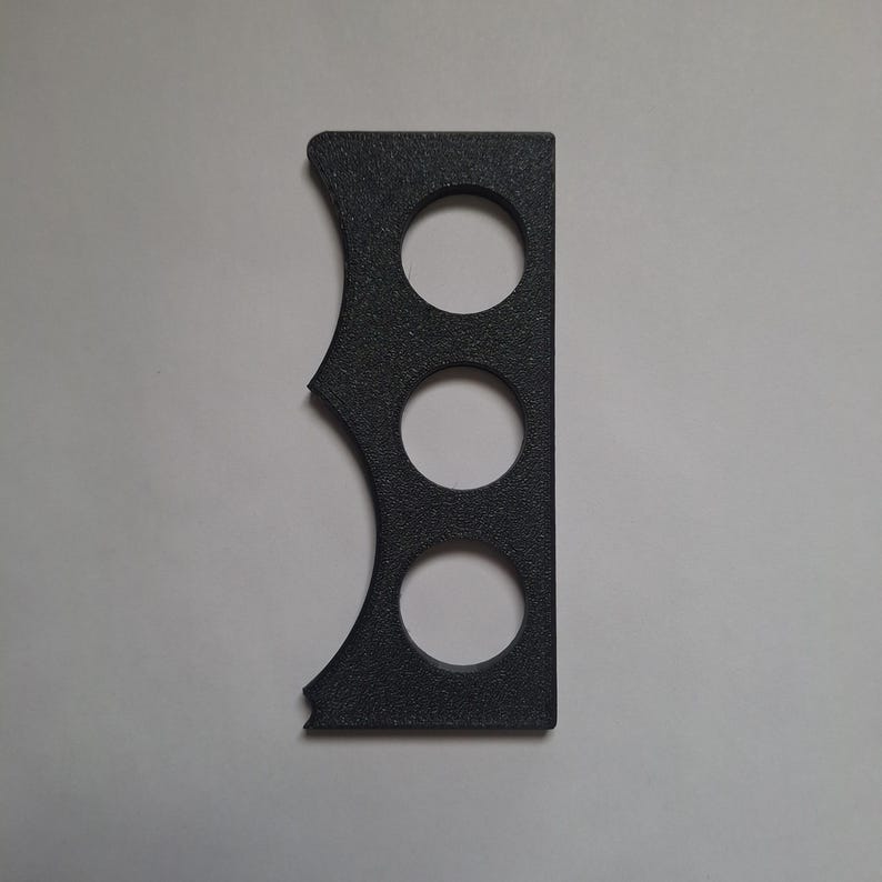 May include: A rectangular black object with a textured surface featuring three circular cutouts arranged vertically in the center and a scalloped edge along one side. The object has a flat, straight edge on the opposite side and a matte finish.
