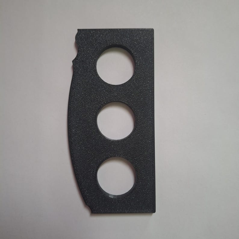 May include: A dark grey, rectangular, textured plastic component featuring three evenly spaced circular cutouts arranged vertically. The left edge has a curved profile with two small notches, while the right edge is straight and flat.