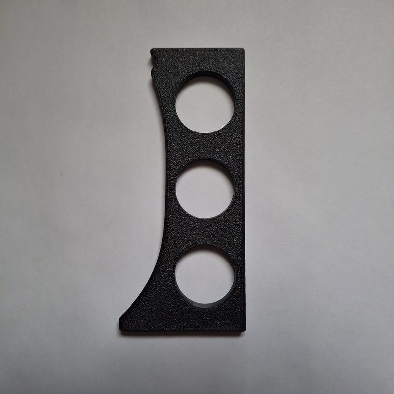 Puede incluir: Un objeto rectangular negro con textura, que presenta un lado izquierdo curvo y tres aberturas circulares dispuestas verticalmente. El art&iacute;culo tiene un acabado mate y granulado, con un borde derecho recto y funcional.