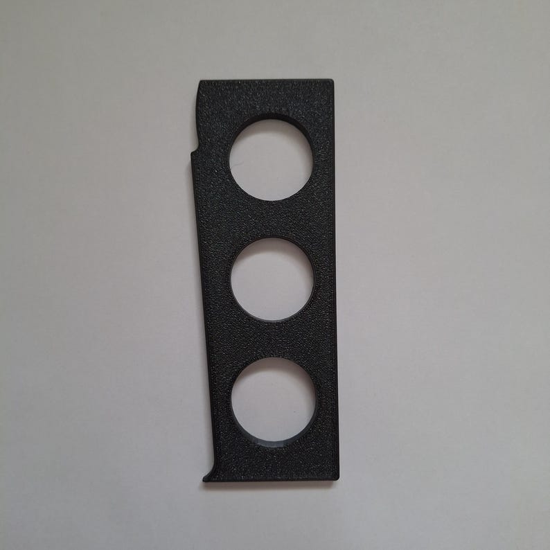 May include: A rectangular black plastic component featuring a textured surface and three evenly spaced circular cutouts arranged vertically. The piece has a straight right edge and a slightly contoured left edge with a small notch near the top.