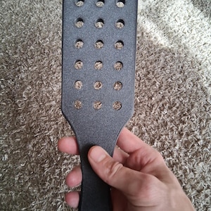 May include: A black paddle with a rectangular head and a handle. The head of the paddle has multiple evenly spaced circular holes. The paddle is held in a hand against a textured, neutral-colored background.