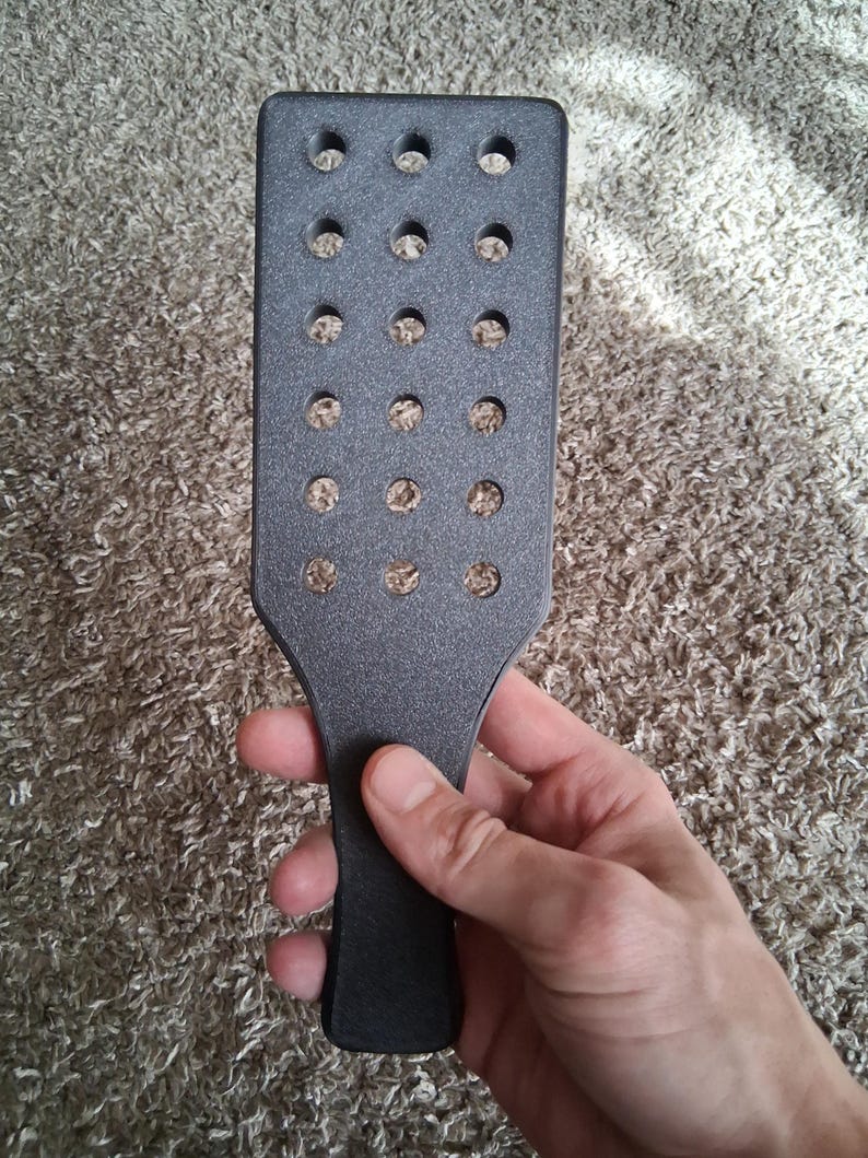 May include: A dark grey paddle with a rectangular head and a handle. The head features numerous evenly spaced circular holes. The paddle is held in a hand against a textured, neutral-toned background.