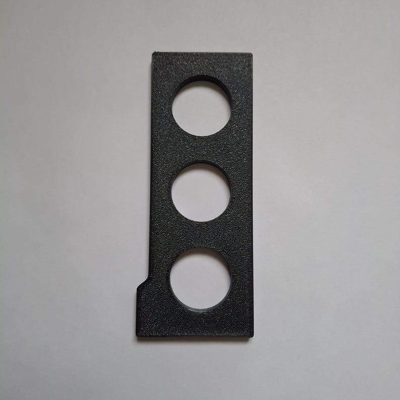 Puede incluir: Un componente rectangular de pl&aacute;stico negro con una superficie texturizada que presenta tres aberturas circulares espaciadas uniformemente dispuestas verticalmente. La pieza tiene un borde derecho recto y una esquina inferior izquierda muescada.