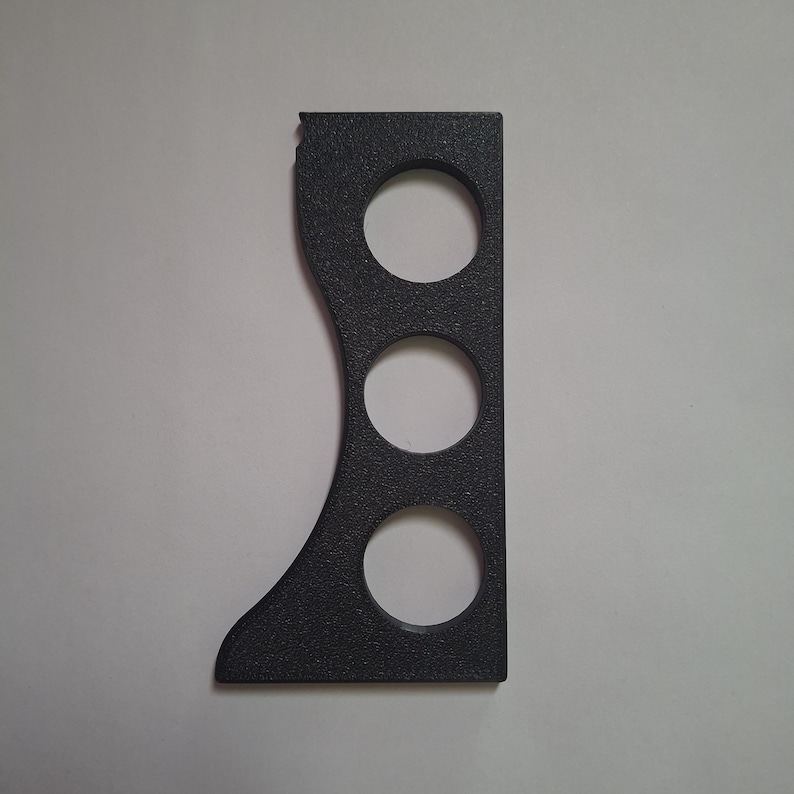 Puede incluir: Un componente negro, texturizado, plano y rectangular que presenta un borde izquierdo curvo y tres recortes circulares dispuestos verticalmente. El objeto tiene un borde derecho recto, una base plana y una peque&ntilde;a muesca en la esquina superior.