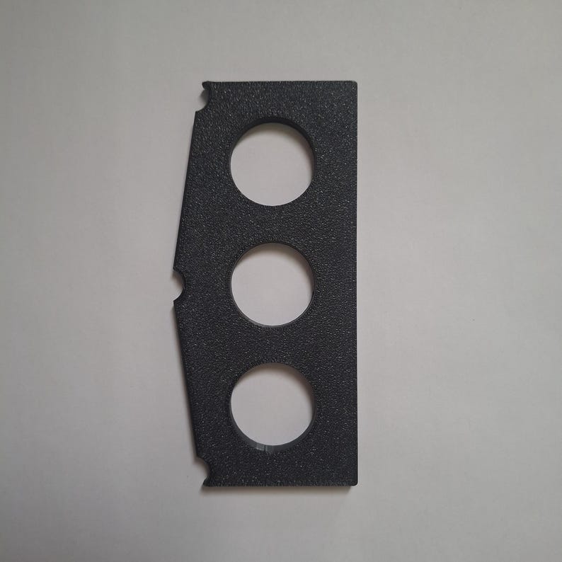 Puede incluir: Un componente de pl&aacute;stico gris oscuro, rectangular y texturizado, que presenta tres recortes circulares dispuestos verticalmente en el centro y dos muescas semicirculares a lo largo del borde izquierdo, con un lado derecho plano y un lado izquierdo inclinado.