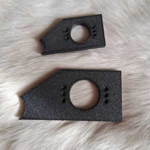 Puede incluir: Dos soportes metálicos negros texturizados. Cada soporte tiene un agujero circular grande y tres agujeros más pequeños. Los soportes tienen una forma única con un borde dentado. El fondo es un material suave, blanco y peludo.