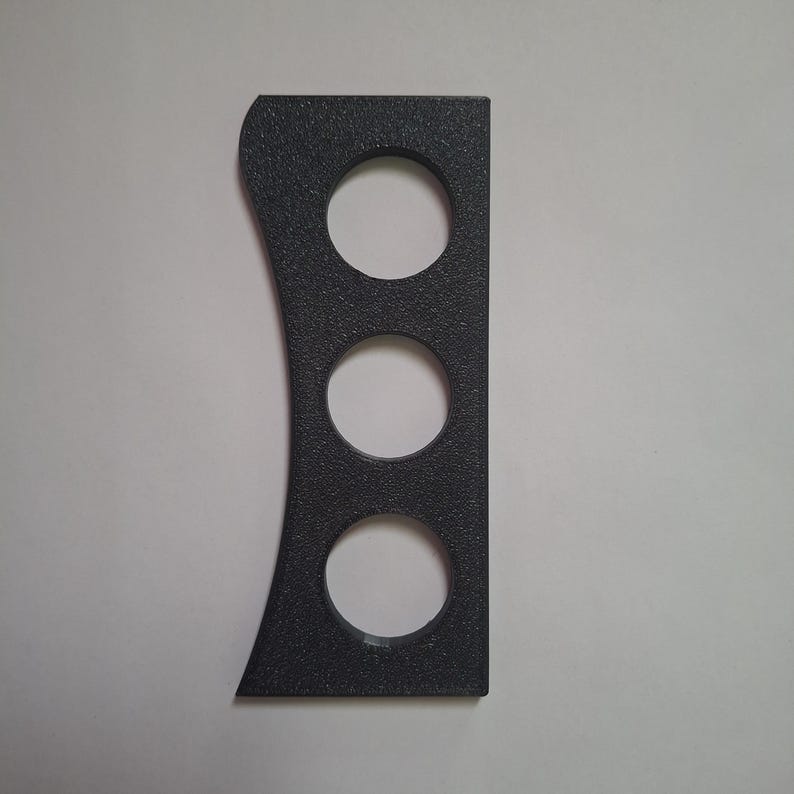Puede incluir: Un objeto rectangular de color gris oscuro con textura, que presenta un borde derecho recto y un borde izquierdo curvo. El objeto cuenta con tres aberturas circulares espaciadas uniformemente y alineadas verticalmente, creando un dise&ntilde;o perforado.