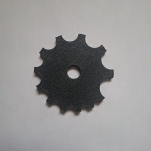 May include: A flat, circular gear-shaped component with a textured, matte black finish. The object features ten evenly spaced rounded teeth along its outer perimeter and a single, centered circular hole for mounting or attachment purposes.