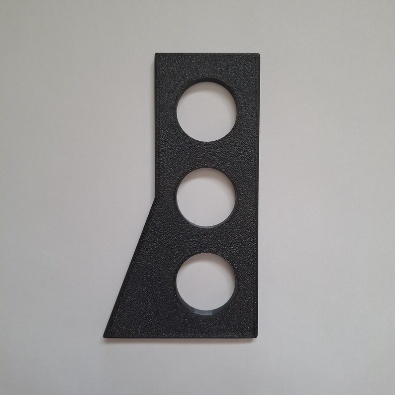 May include: A dark gray, textured, flat, rigid component with a vertical rectangular shape on one side and an angled edge on the other. It features three evenly spaced circular cutouts arranged in a vertical line along the center of the piece.