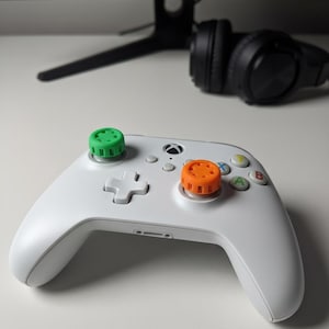 May include: A white Xbox controller with green and orange thumbstick covers. The controller has a directional pad, buttons, and a central Xbox button. Black headphones and a computer monitor are in the background.